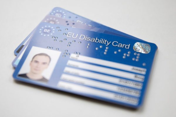 An example of the Belgian version of the European Disability Card - a blue card with some white fileds for personal information, a rectangular space for a photo and a silver chip. The Card has braille embossed