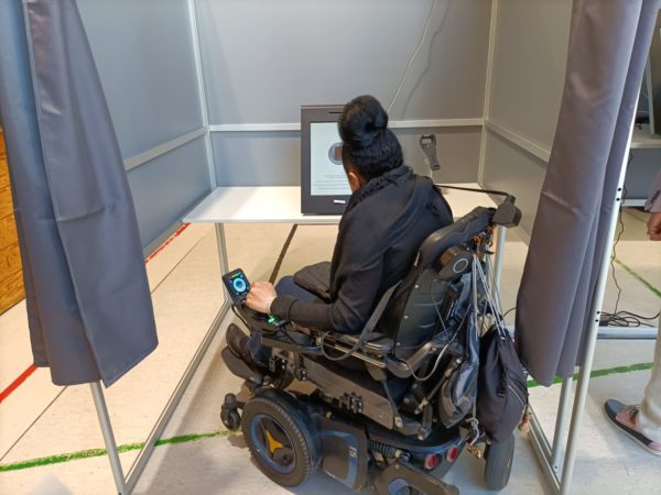 Nadia in the adapted voting booth, which is clearly too smal for her and her wheelchair and we can notice that the curtains don't even reach half of the need space to close around her